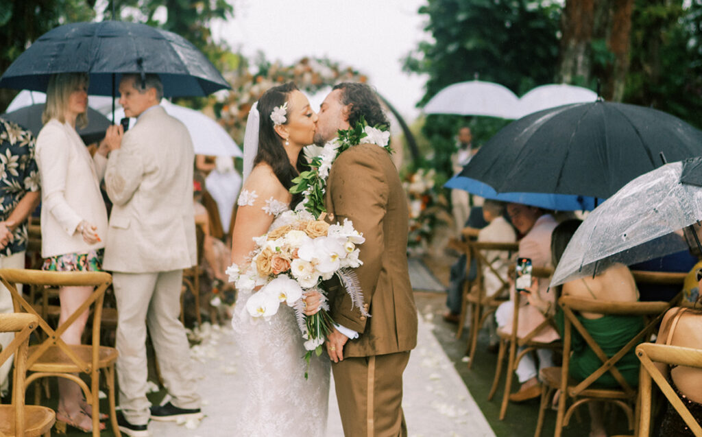 Bride and groom kissing under clear umbrellas during a rainy Hawaii wedding ceremony.