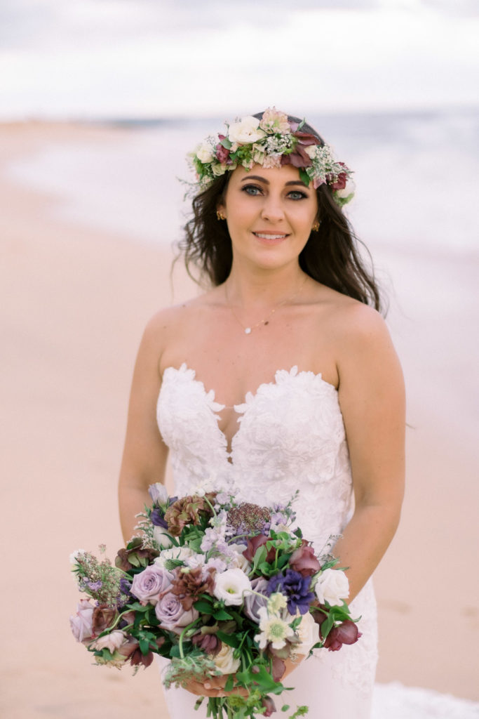 The bride in her wedding dress on the beach with a bouquet and flowers in her hair.