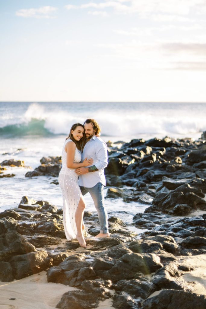 A woman with brown hair and a white body con dress holds her boyfriend while standing on black lava rocks in Kauai Hawaii.