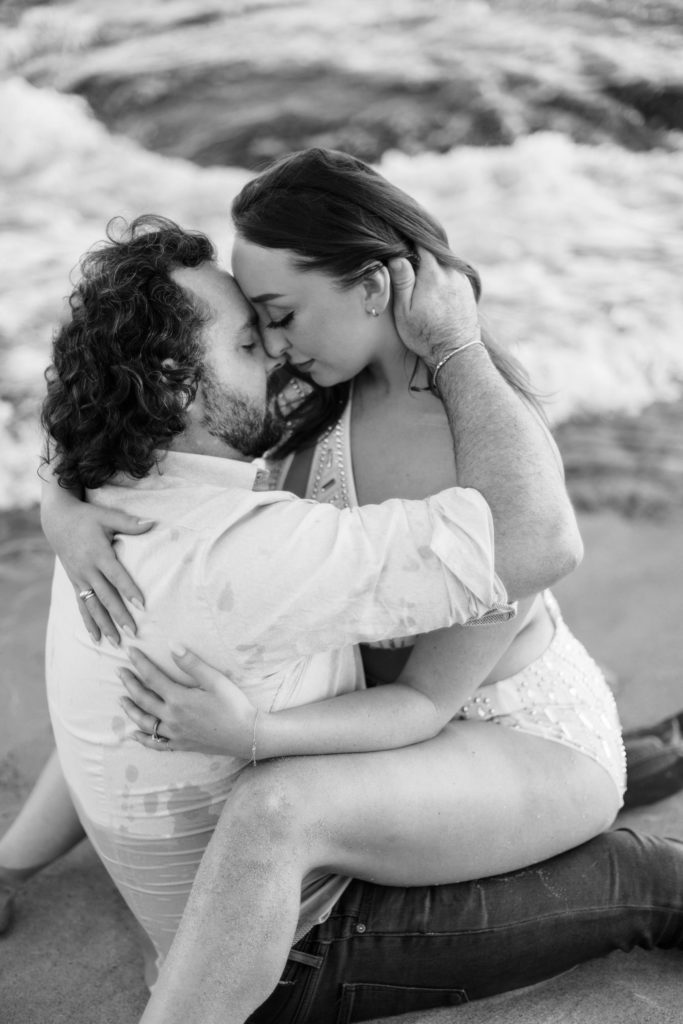 A man and woman embrace while sitting down in the water on the sand.  