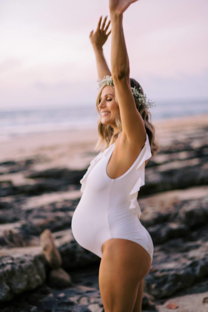 smiling young pregnant mother with her hands in the air and wearing a white swim suit.
