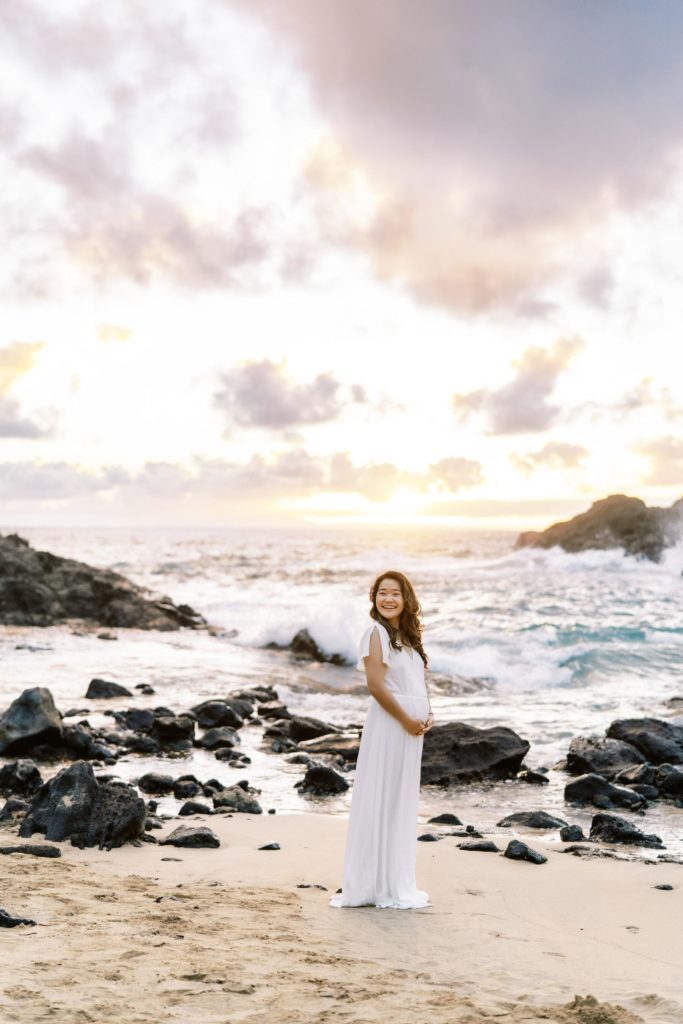 A pregnant woman standing on a beach in hawaii in a white maternity dress.