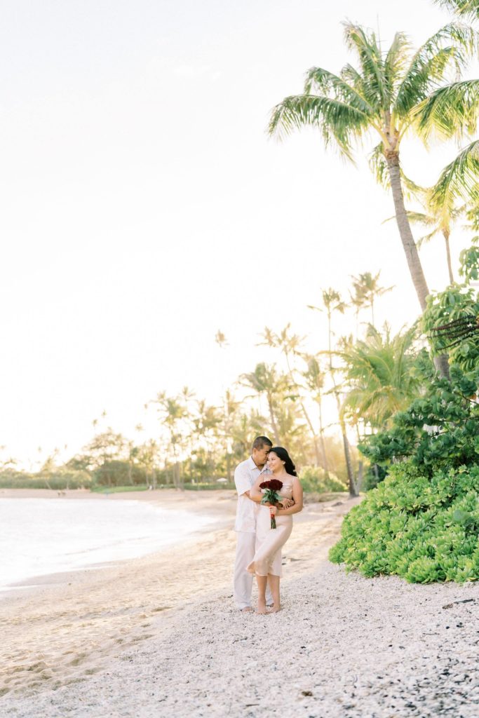 man holding his fiance with his arms around her waist.  Coconut trees along a white sand beach. 