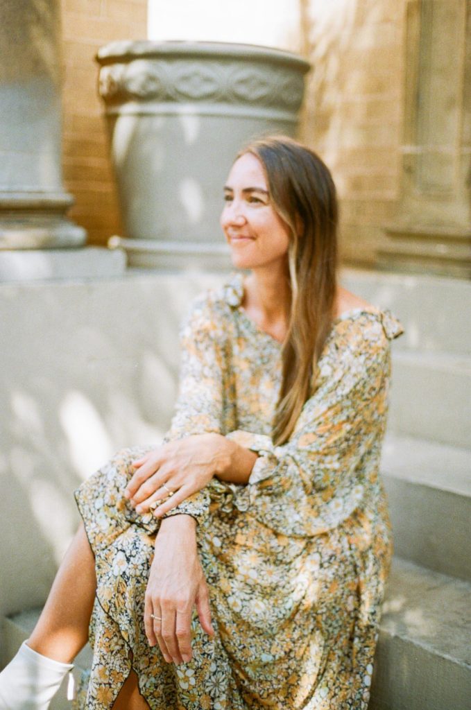 Blur photo of a woman in floral dress and white boots sitting on the steps smiling and looking away 