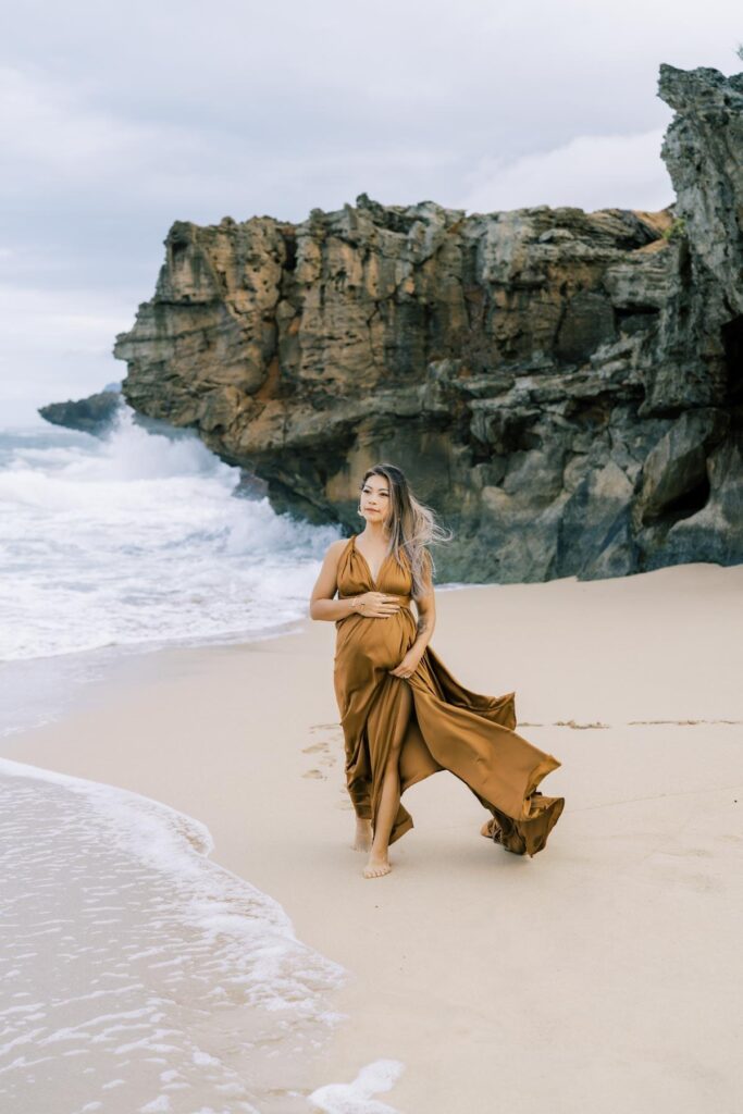Pregnant woman holding her baby bump wearing a brown dress walking towards the beach