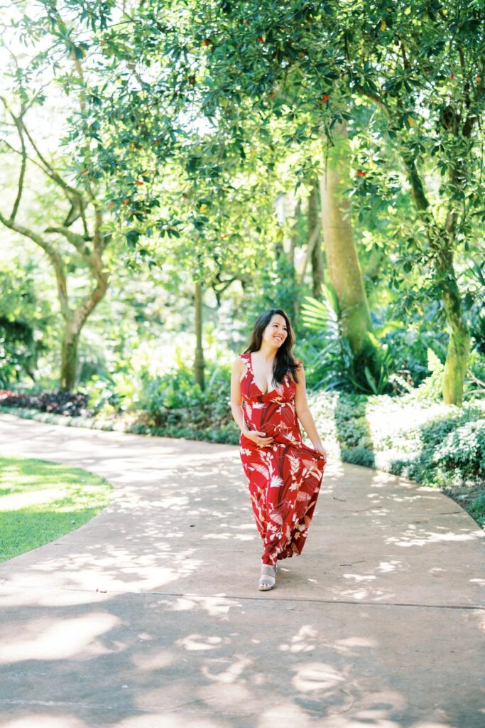 A Pregnant woman walking wearing a long red floral dress on her Pregnancy Photo Session at Oahu Botanical Garden