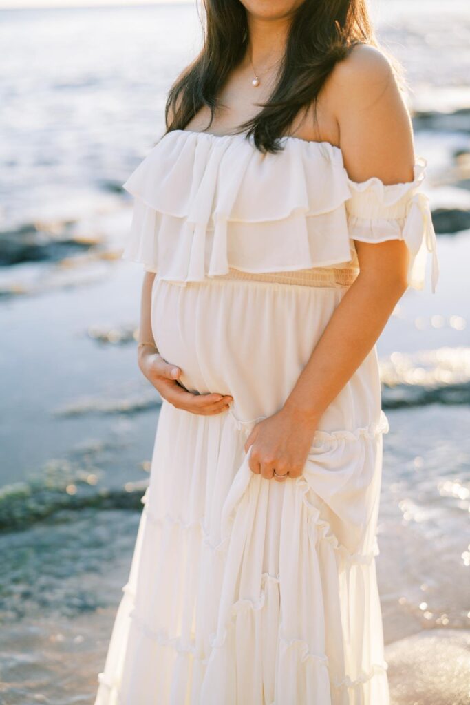 A Pregnant woman wearing an off-shoulder white dress, holding her baby bump on her Pregnancy photo Session at the beach