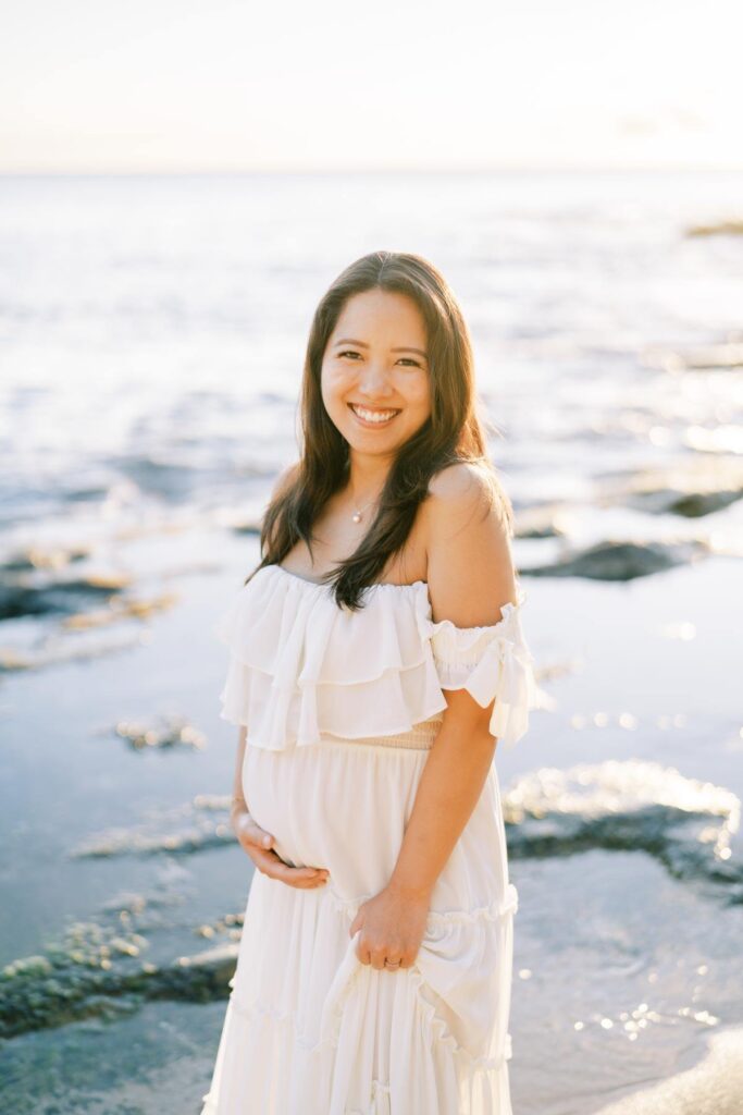 A Pregnant woman wearing an off-shoulder white dress, smiling, holding her baby bump on her Pregnancy photo Session at the beach