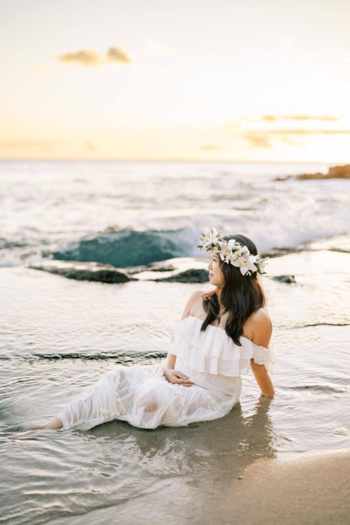 A Pregnant woman wearing an off-shoulder white dress, holding her baby bump on her Pregnancy photo Session at the beach