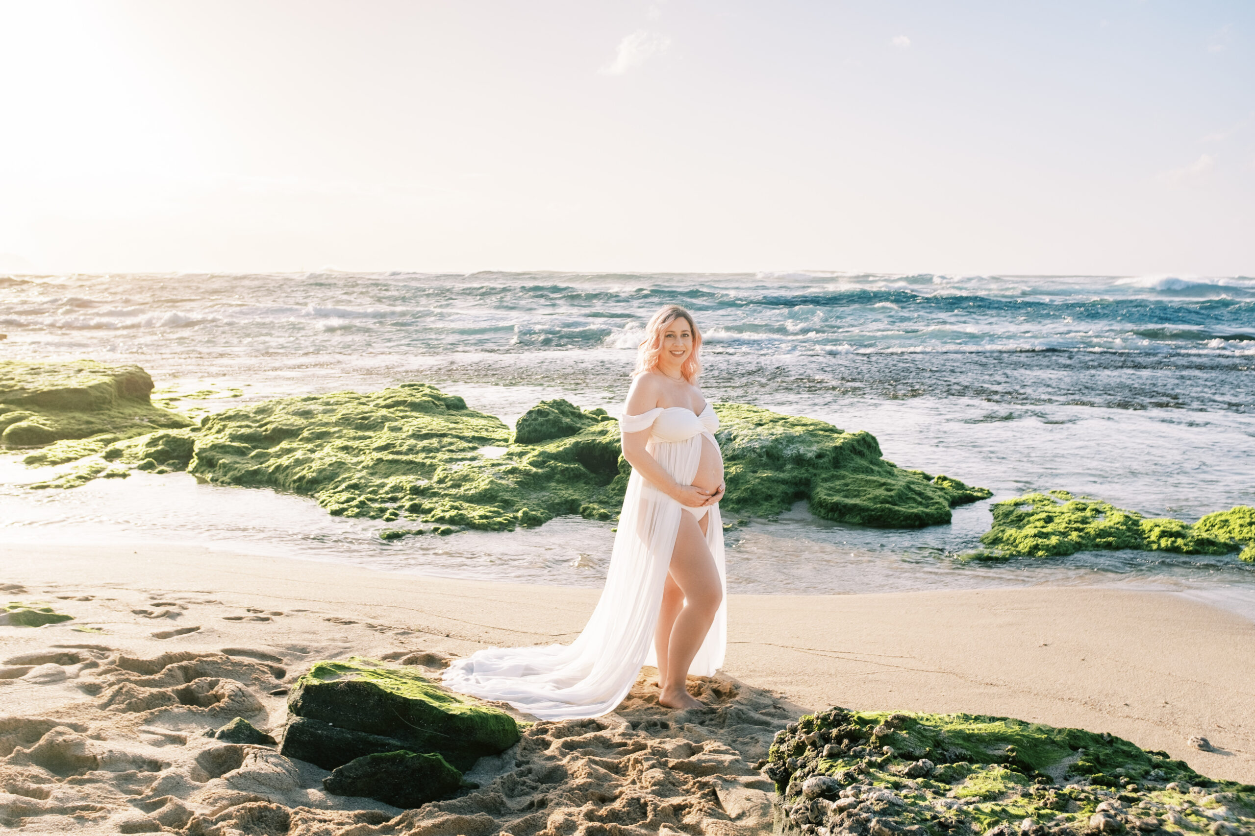 Woman in a flowing dress walking on a moss-covered rock by the sea at sunset during their maternity session on Oahu.