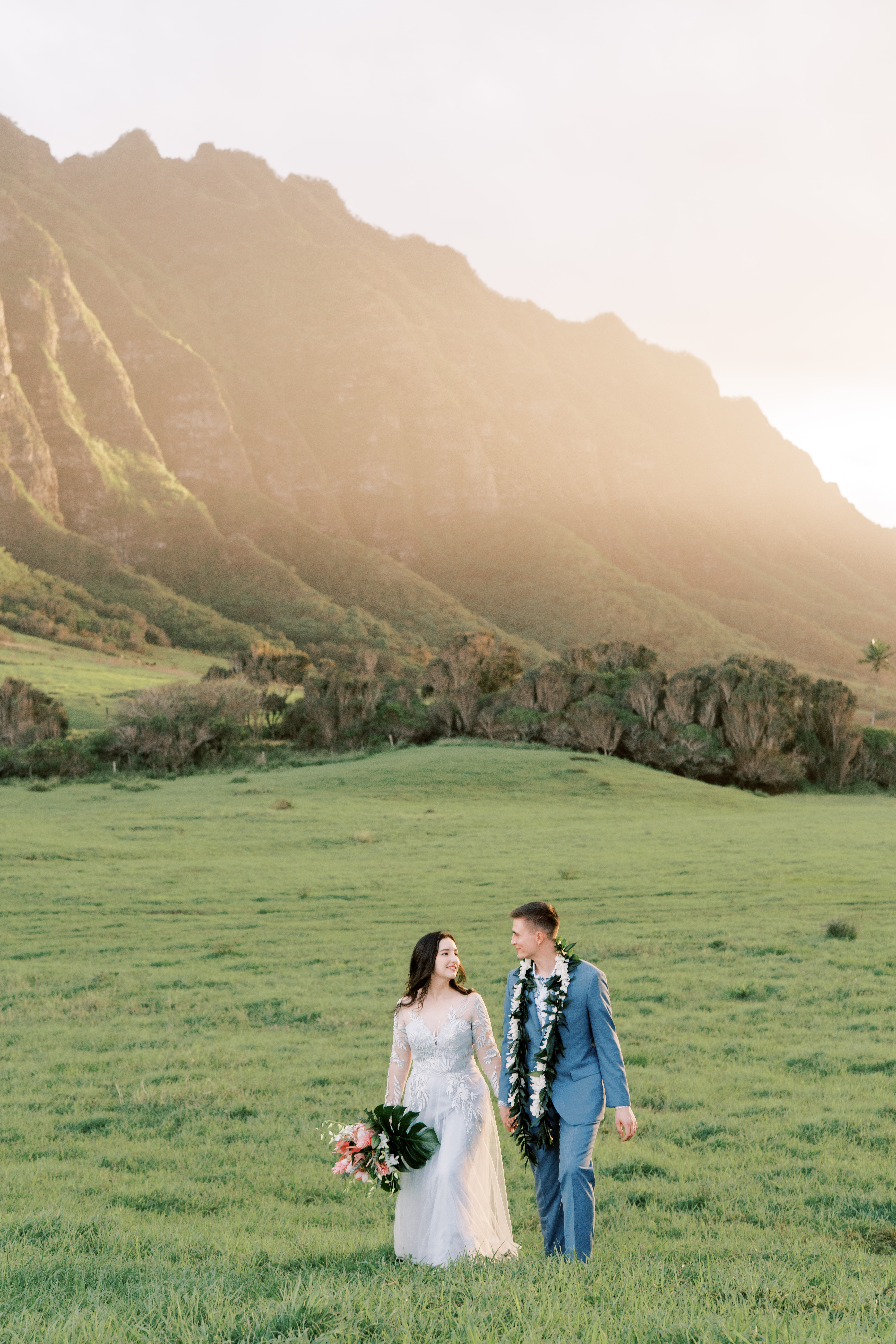 A bride and groom walking through a grassy field at Kualoa Ranch, with mountains in the background.
