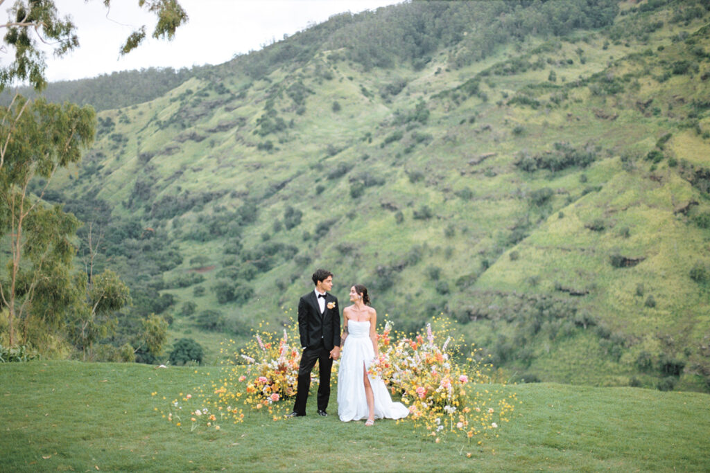 couple in wedding attire, standing together on a hill with flowers