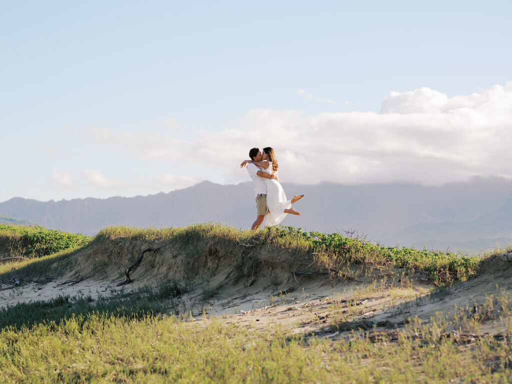 Newly engaged couple. Man is lifting up his fiance and twirling. Mountains in the background and green grass. Private Beach Oahu Engagement