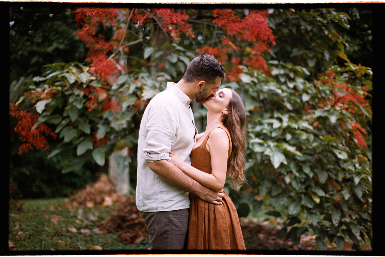 Couple kissing during an engagement session at Ho’omaluhia Botanical Garden surrounded by lush greenery and red tropical flowers.
